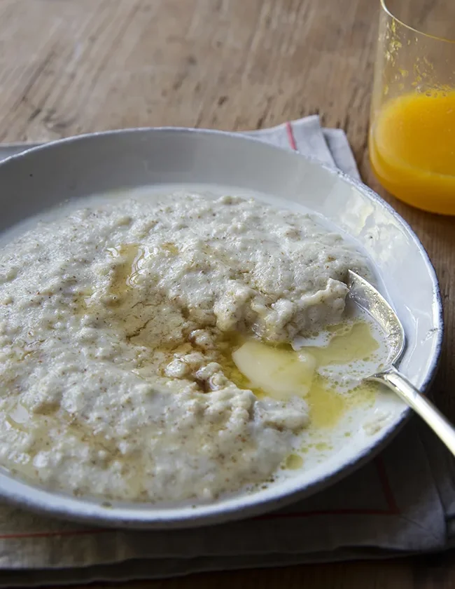 Ina Garten maple vanilla cream of wheat in a gray bowl with melting butter and maple syrup, next to a glass of orange juice