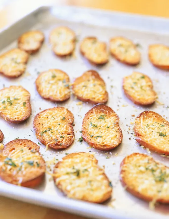 Ina Garten parmesan croutons, golden baguette slices topped with melted shredded Parmesan and herbs on a baking sheet