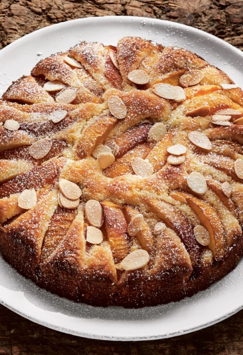 Ina Garten peach almond torte topped with concentric rings of golden peach wedges, sliced almonds and confectioners' sugar on a white plate
