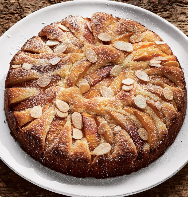 Ina Garten peach almond torte topped with concentric rings of golden peach wedges, sliced almonds and confectioners' sugar on a white plate