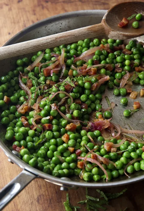 Ina Garten peas & pancetta in a stainless steel sauté pan with sliced shallot, diced pancetta and fresh mint, with a wooden spoon