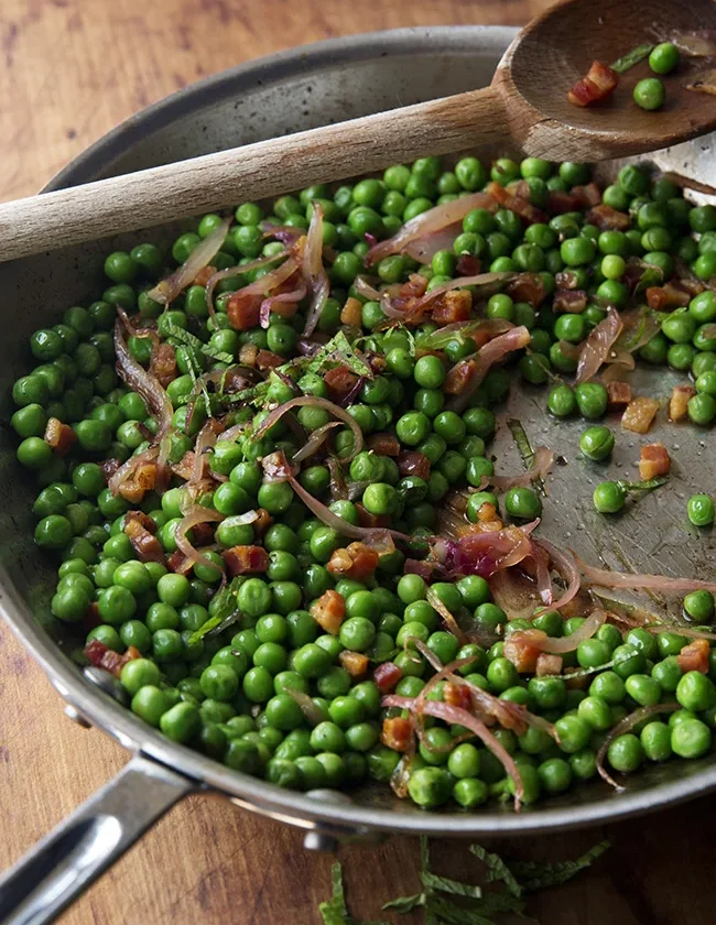 Ina Garten peas & pancetta in a stainless steel sauté pan with sliced shallot, diced pancetta and fresh mint, with a wooden spoon