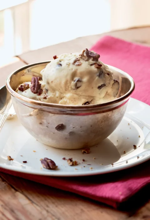 Ina Garten pecan rum raisin ice cream scoop in a silver bowl topped with chopped pecans on a pink napkin