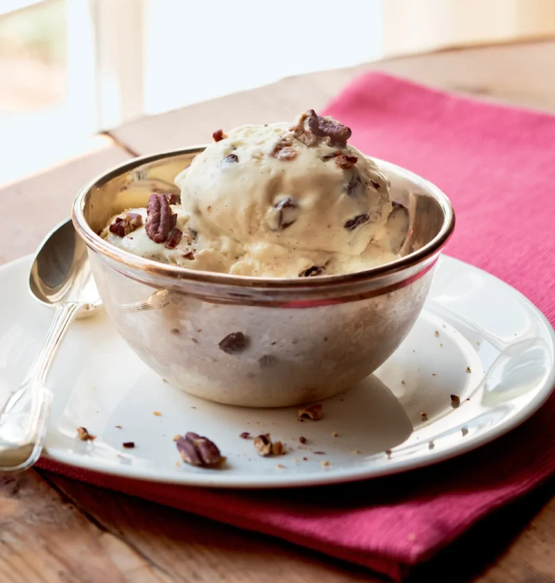 Ina Garten pecan rum raisin ice cream scoop in a silver bowl topped with chopped pecans on a pink napkin