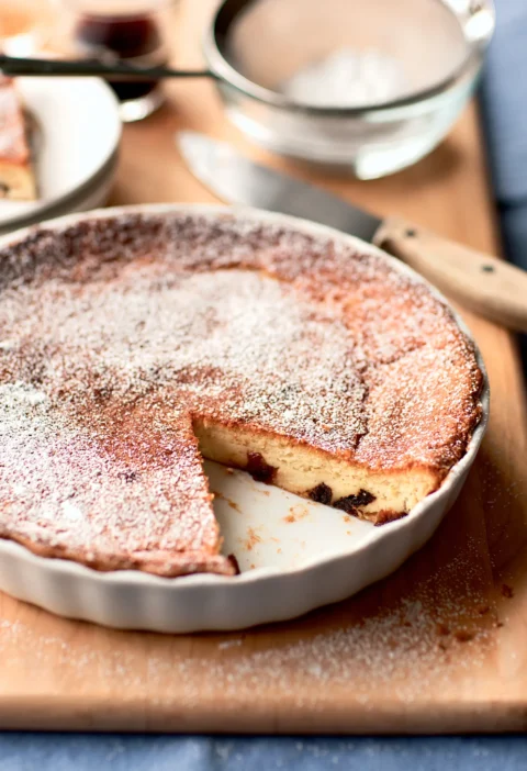 Ina Garten prune Armagnac clafouti dusted with confectioners' sugar in a white baking dish with a slice cut out