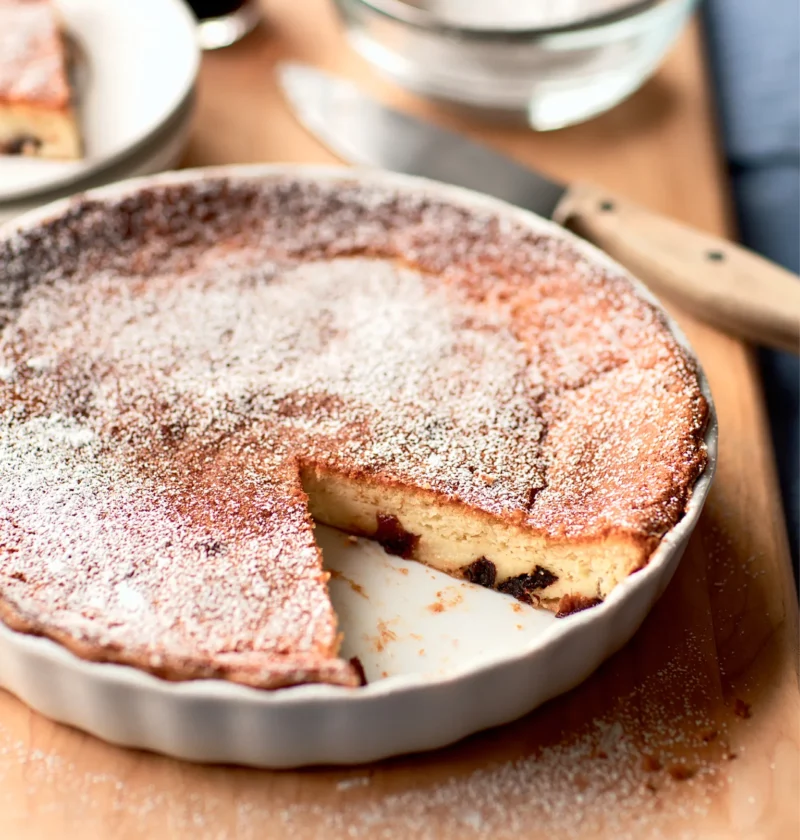 Ina Garten prune Armagnac clafouti dusted with confectioners' sugar in a white baking dish with a slice cut out