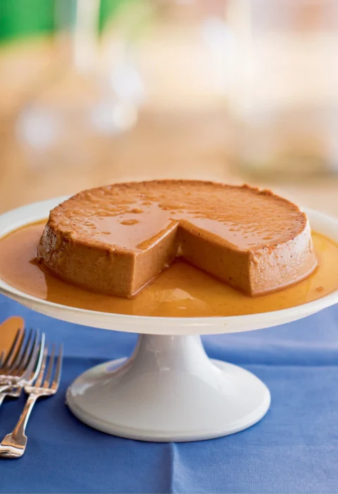 Ina Garten pumpkin flan with maple caramel on a white cake stand with a wedge cut out and caramel pooling around it