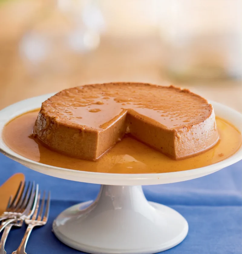 Ina Garten pumpkin flan with maple caramel on a white cake stand with a wedge cut out and caramel pooling around it