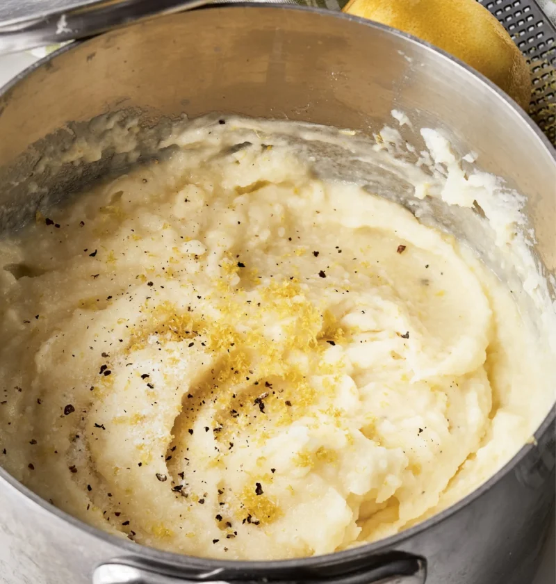 Ina Garten puréed potatoes with lemon in a saucepan, topped with lemon zest and black pepper, with a lemon and grater beside it