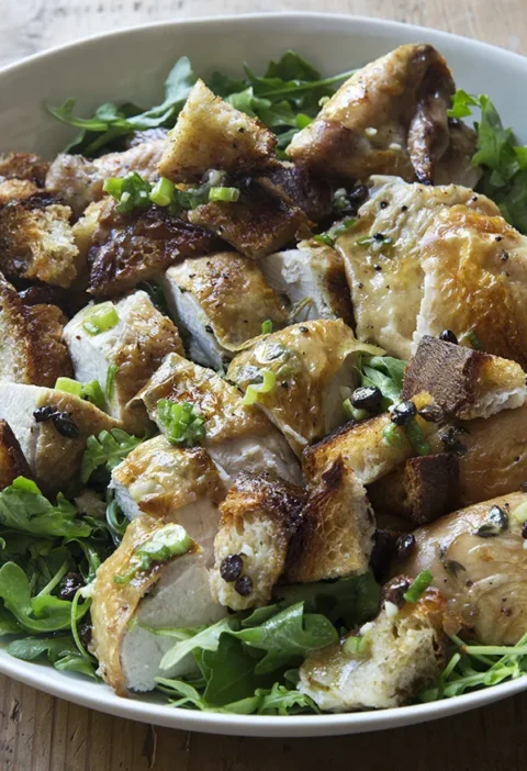 Ina Garten roast chicken with bread and arugula salad, carved chicken and toasted bread squares on a white platter of arugula
