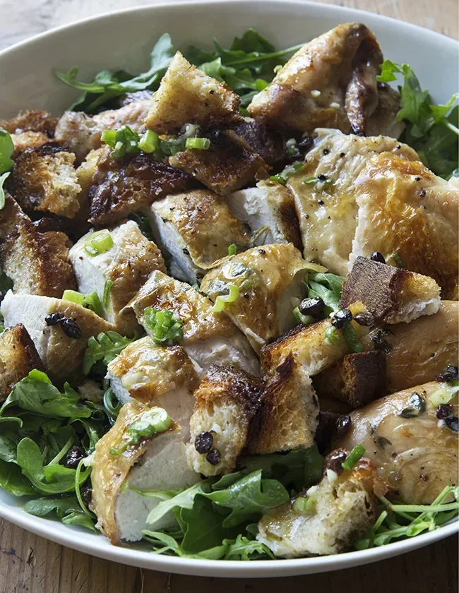 Ina Garten roast chicken with bread and arugula salad, carved chicken and toasted bread squares on a white platter of arugula