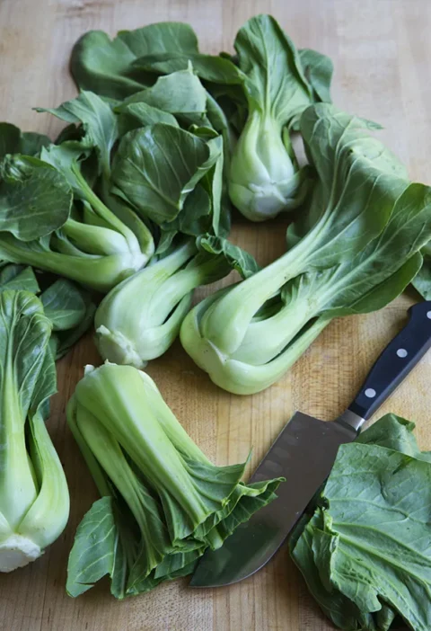 Fresh baby bok choy heads piled on a wooden cutting board next to a chef's knife, ready for Ina Garten roasted baby bok choy