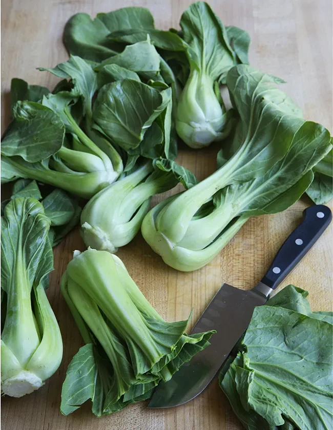 Fresh baby bok choy heads piled on a wooden cutting board next to a chef's knife, ready for Ina Garten roasted baby bok choy