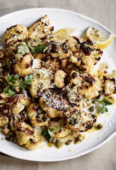 Ina Garten roasted cauliflower with lemon & capers on a white platter with Parmesan, parsley and lemon wedges