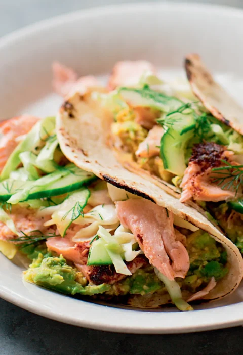 Ina Garten roasted salmon tacos with avocado, cucumber and cabbage slaw with dill in corn tortillas on a white plate