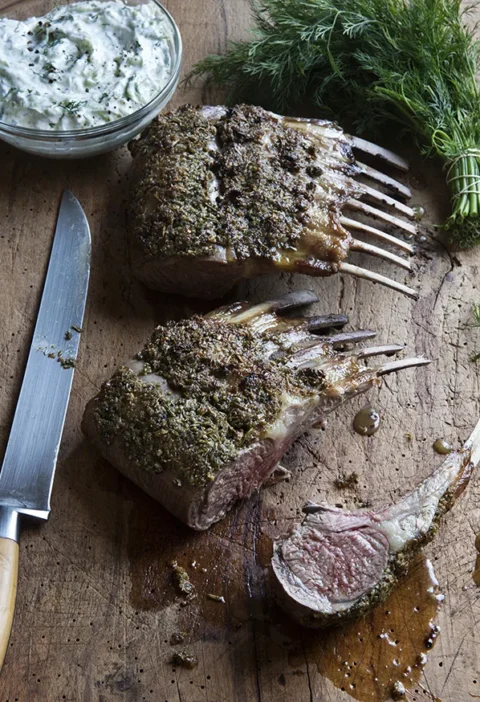 Ina Garten rosemary rack of lamb with herb crust sliced into chops on a wooden board next to a bowl of easy tzatziki and fresh dill