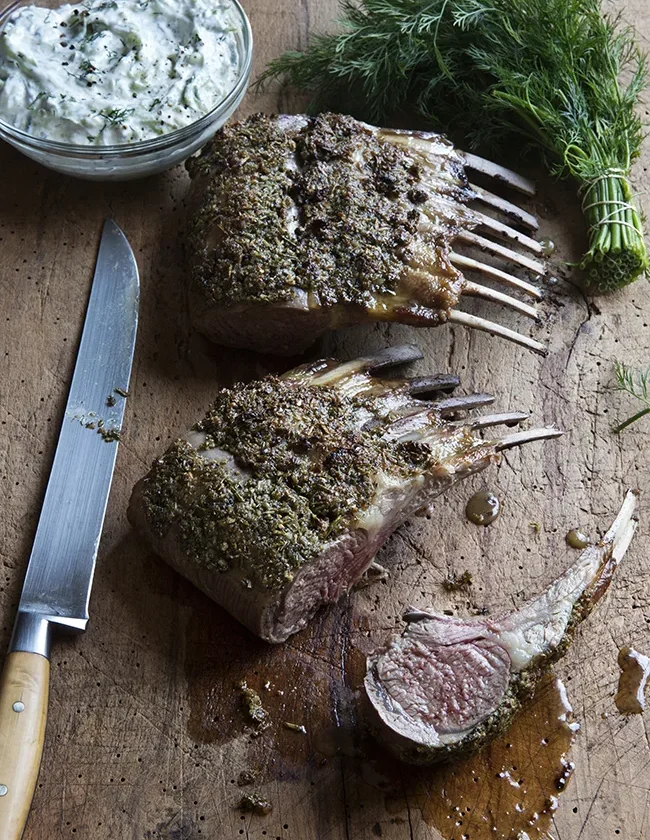 Ina Garten rosemary rack of lamb with herb crust sliced into chops on a wooden board next to a bowl of easy tzatziki and fresh dill