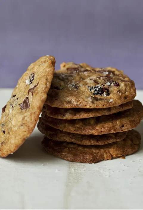 Stack of salty oatmeal chocolate chunk cookies with dried cranberries and flaky salt, with one cookie leaning on the side