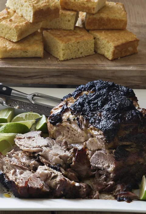 Ina Garten slow-roasted spiced pork with a dark crackly crust, sliced and served with lime wedges and corn bread squares behind