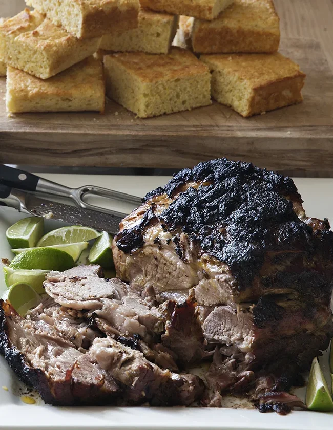 Ina Garten slow-roasted spiced pork with a dark crackly crust, sliced and served with lime wedges and corn bread squares behind