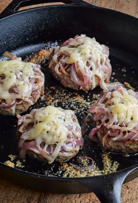 Ina Garten smashed hamburgers with caramelized onions and melted Gruyère cooking in a cast-iron skillet