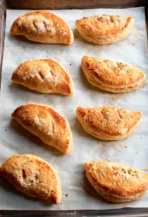 Ina Garten spicy sweet potato empanadas, golden puff pastry half circles sprinkled with flaked sea salt on a parchment-lined sheet pan