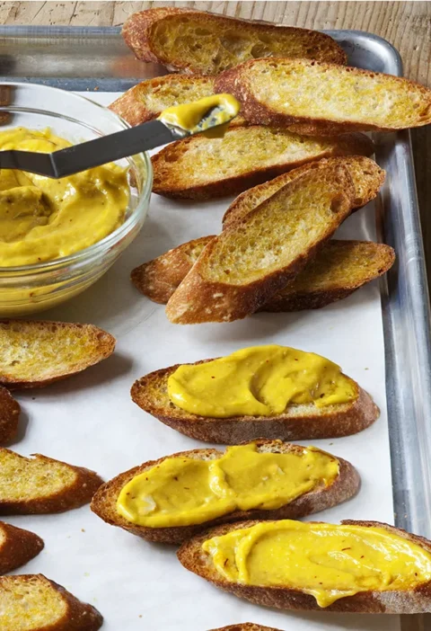 Ina Garten Sriracha rouille spread on toasted baguette slices with a bowl of golden rouille on a baking sheet