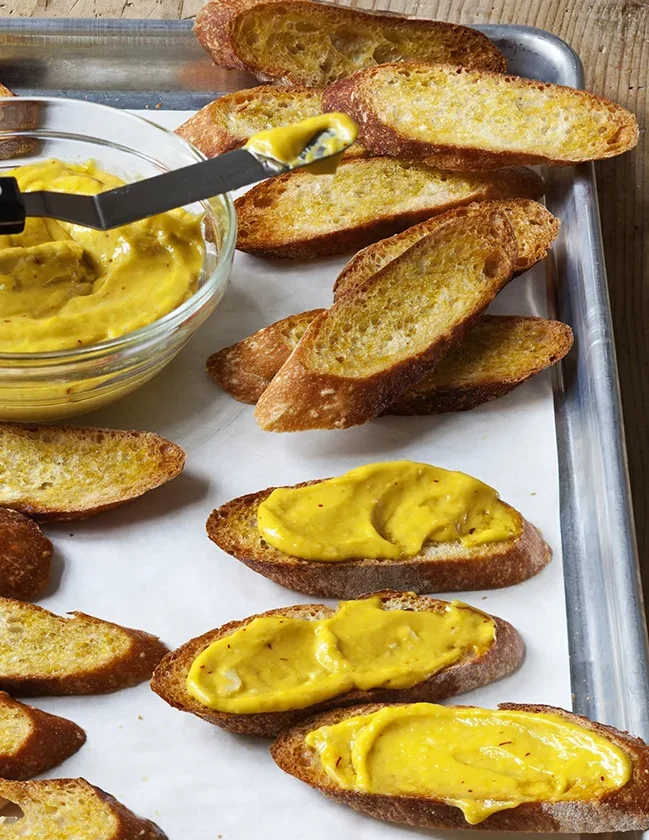 Ina Garten Sriracha rouille spread on toasted baguette slices with a bowl of golden rouille on a baking sheet