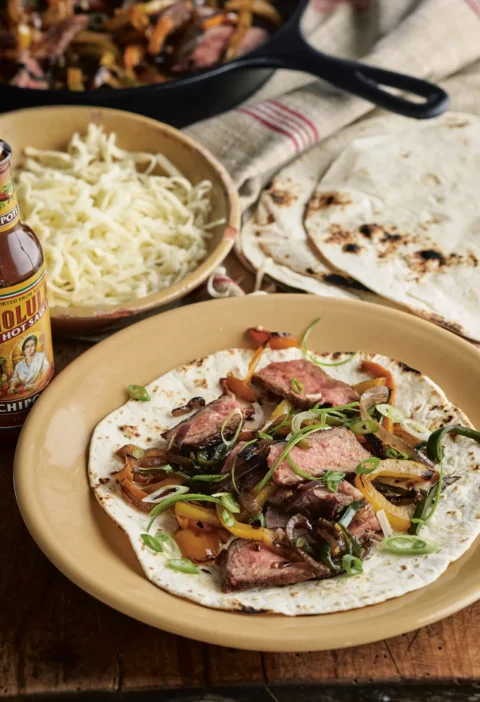 Ina Garten steak fajitas with sliced steak, blistered peppers and scallions on a flour tortilla beside a bowl of shredded cheese