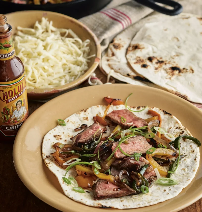 Ina Garten steak fajitas with sliced steak, blistered peppers and scallions on a flour tortilla beside a bowl of shredded cheese