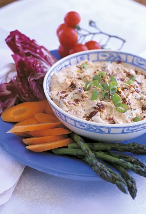 Ina Garten sun-dried tomato dip in a blue and white bowl topped with sliced scallions, served with carrots, asparagus and radicchio