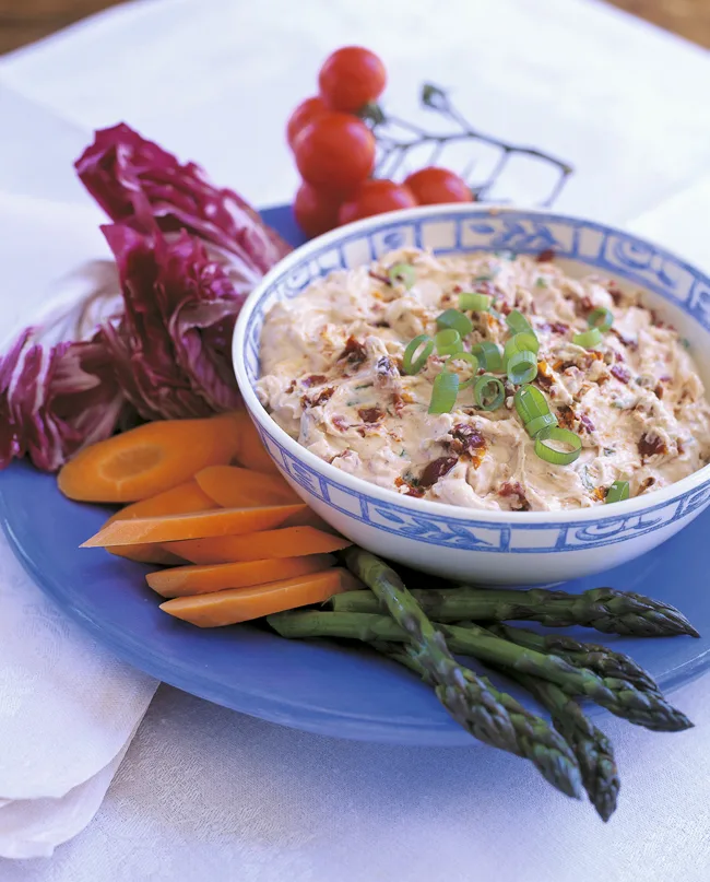 Ina Garten sun-dried tomato dip in a blue and white bowl topped with sliced scallions, served with carrots, asparagus and radicchio