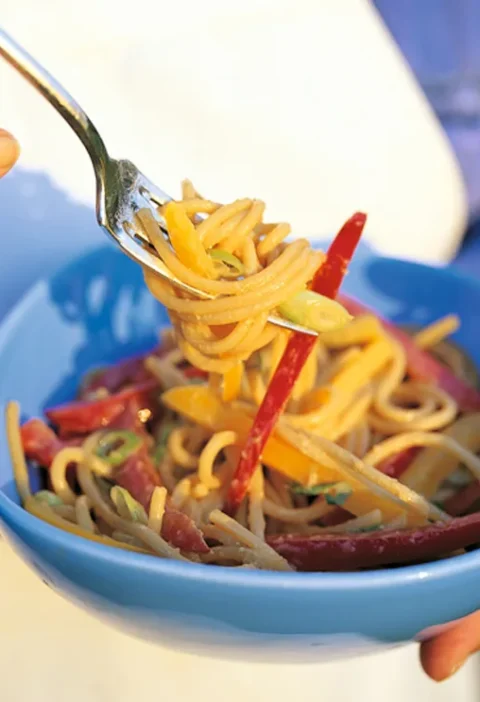 Ina Garten Szechuan noodles twirled on a fork above a blue bowl with red and yellow bell pepper strips and sliced scallions