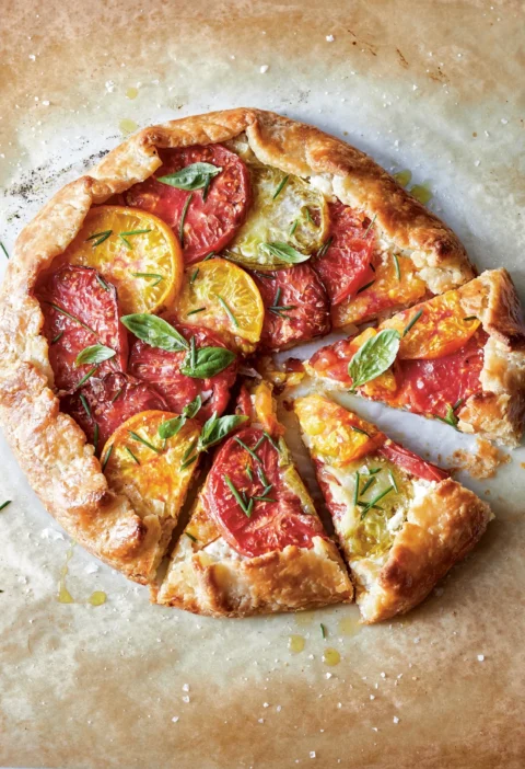 Ina Garten tomato & goat cheese crostata topped with red and yellow heirloom tomato slices and fresh basil on parchment paper