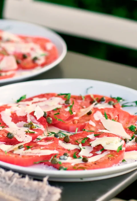Ina Garten tomato carpaccio with overlapping tomato slices topped with capers, basil and shaved Parmesan on a white plate