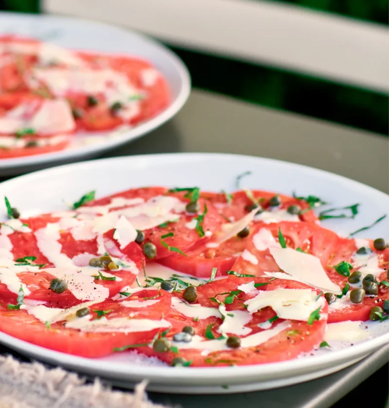 Ina Garten tomato carpaccio with overlapping tomato slices topped with capers, basil and shaved Parmesan on a white plate