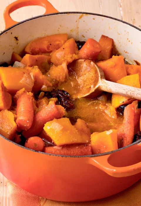 Ina Garten tsimmes with carrots, sweet potatoes, butternut squash and prunes in an orange Dutch oven with a wooden spoon