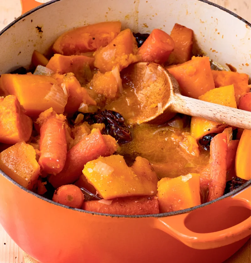 Ina Garten tsimmes with carrots, sweet potatoes, butternut squash and prunes in an orange Dutch oven with a wooden spoon