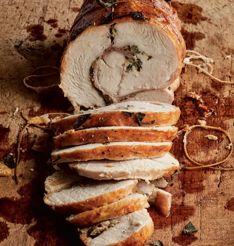 Sliced Ina Garten Tuscan turkey roulade with golden skin, prosciutto, sage and herb filling on a wooden cutting board with kitchen twine
