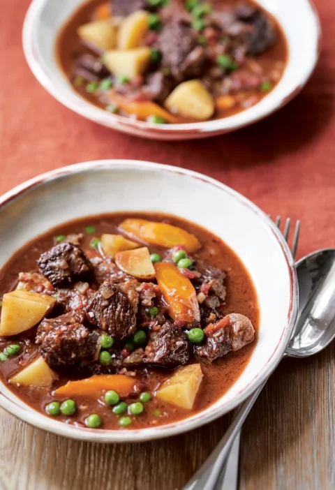 Ina Garten ultimate beef stew with short rib chunks, carrots, potatoes and peas in a white bowl beside a spoon and fork
