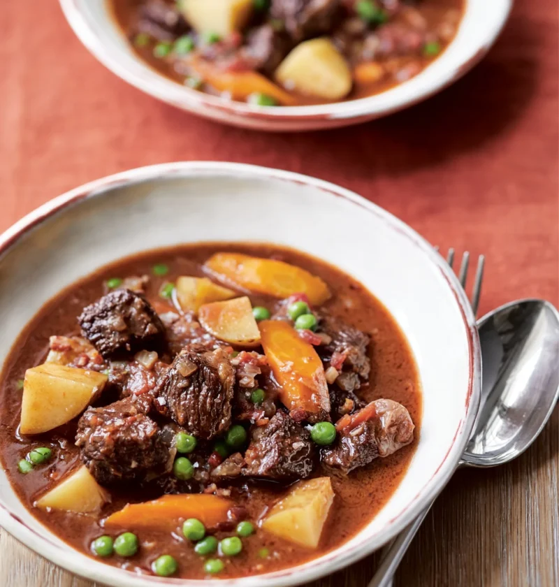 Ina Garten ultimate beef stew with short rib chunks, carrots, potatoes and peas in a white bowl beside a spoon and fork
