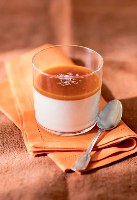 Ina Garten vanilla rum panna cotta with salted caramel in a glass topped with flaky salt on an orange napkin with a spoon