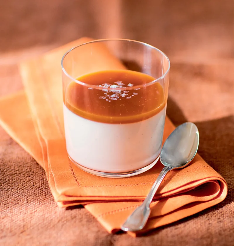 Ina Garten vanilla rum panna cotta with salted caramel in a glass topped with flaky salt on an orange napkin with a spoon