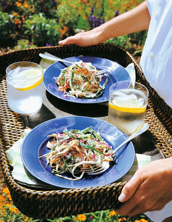 Two blue plates of Ina Garten vegetable coleslaw with red and white cabbage and carrots on a wicker tray beside two glasses of lemon water