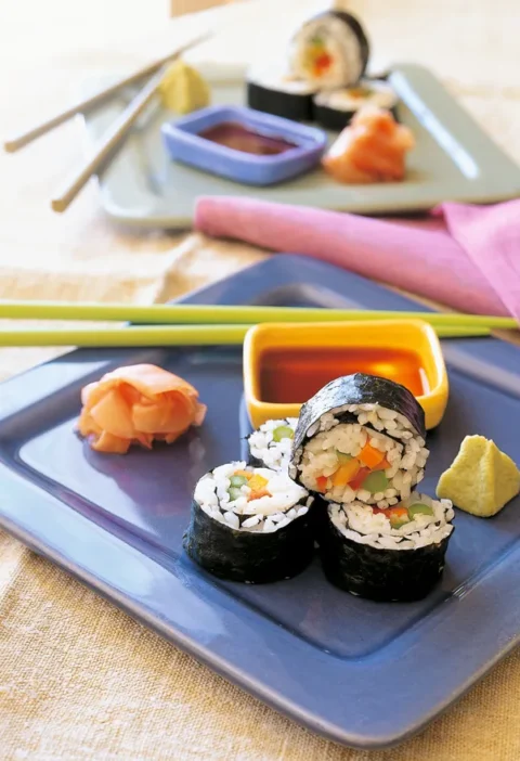 Ina Garten vegetable sushi rolls sliced and stacked on a blue plate with pickled ginger, wasabi and a bowl of dipping sauce