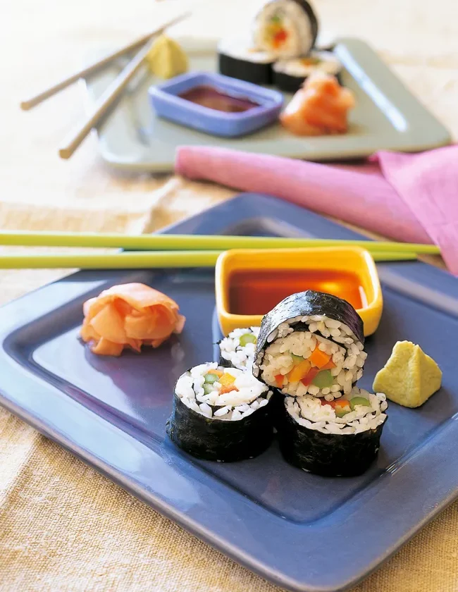 Ina Garten vegetable sushi rolls sliced and stacked on a blue plate with pickled ginger, wasabi and a bowl of dipping sauce