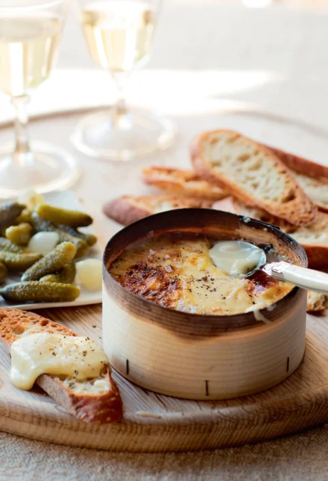 Ina Garten warm vacherin in its wooden box with a spoon, served with toasted bread, cornichons and white wine
